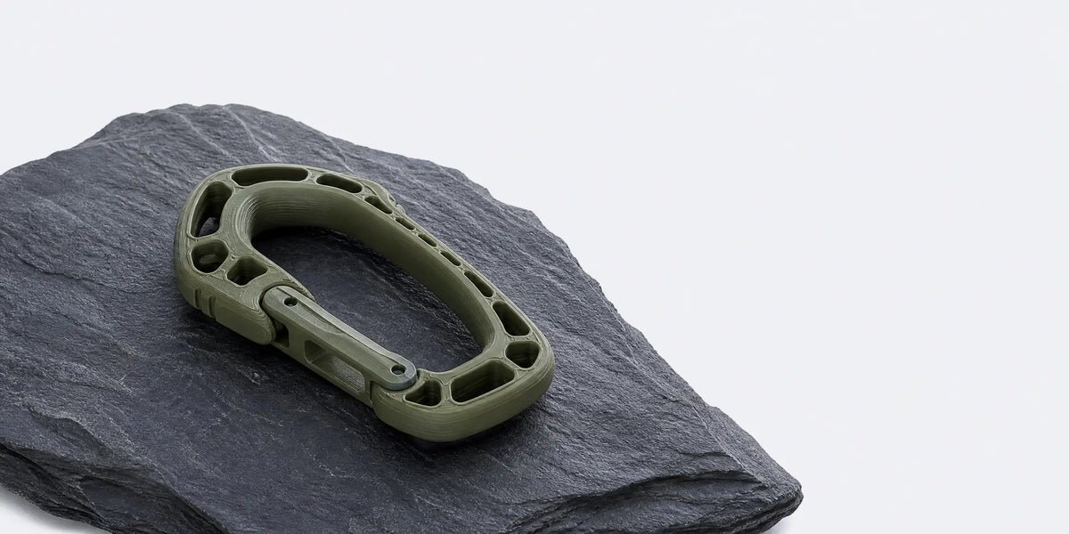 Olive green 3D printed tactical carabiner resting on rough slate stone, highlighting the strength and durability of LANDU filament for outdoor accessories.